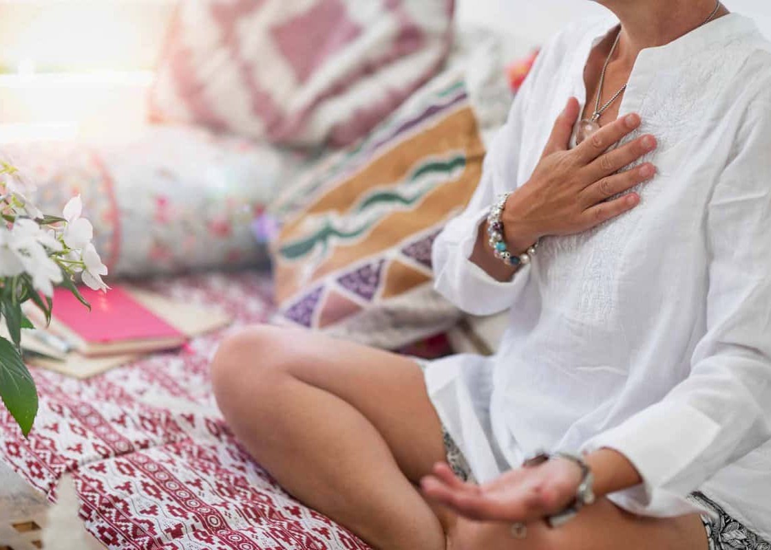 Self-Healing Heart Chakra Meditation. Woman sitting in a lotus position with right hand on heart chakra and left palm open in a receiving gesture. Self-Care Practice at Home