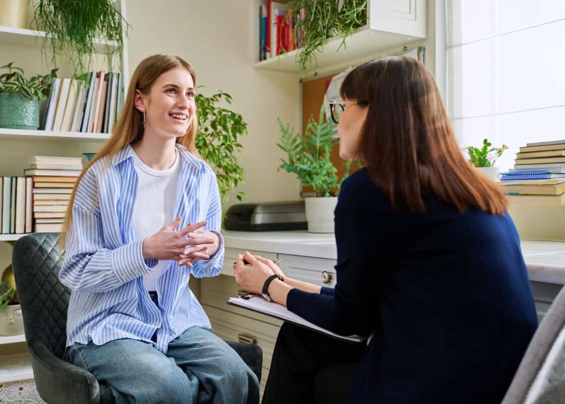 Smiling positive young teen female patient talking to professional mental therapist. Teenager girl high school college student in therapy with psychologist psychotherapist counselor. Treatment support
