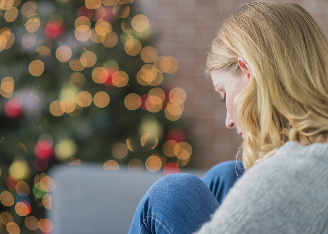 A Caucasian woman is indoors in her living room. There is a Christmas tree in the background. The woman is wearing warm clothing. She is sitting on the couch and looking sad because she is alone on Christmas day.