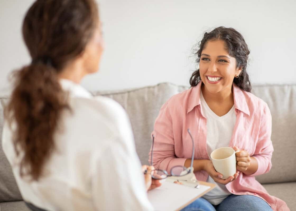 A woman wearing a pink shirt sits on a couch and smiles while holding a cup of coffee. She is talking to another woman sitting next to her, who is wearing a white shirt.