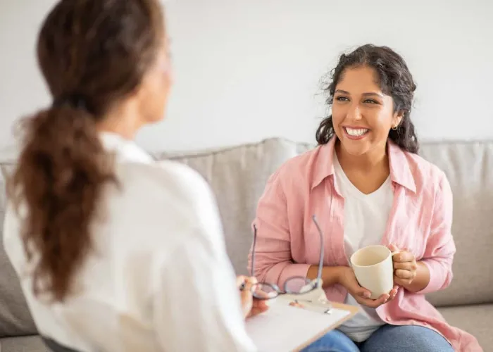 A woman wearing a pink shirt sits on a couch and smiles while holding a cup of coffee. She is talking to another woman sitting next to her, who is wearing a white shirt.