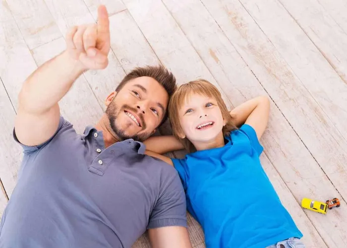 Look over there! Top view of happy father and son lying on the hardwood floor together while young man pointing away and smiling