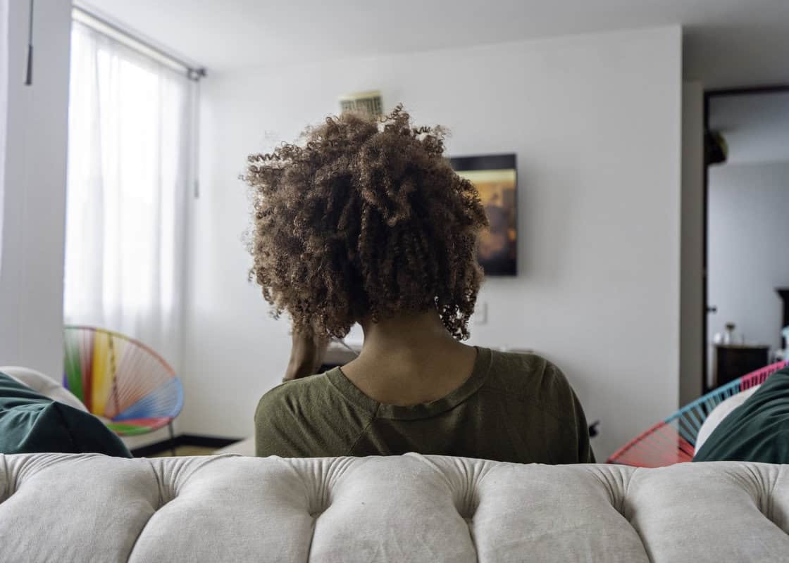 View from the back of an African American woman is watching TV in the living room sitting on the couch in her apartment.