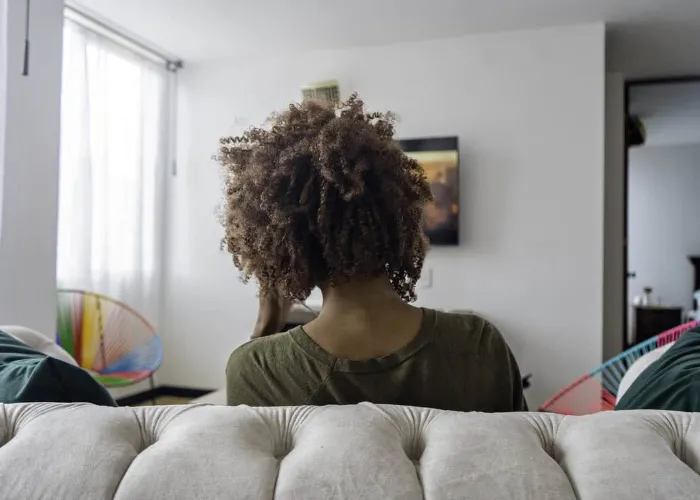 View from the back of an African American woman is watching TV in the living room sitting on the couch in her apartment.