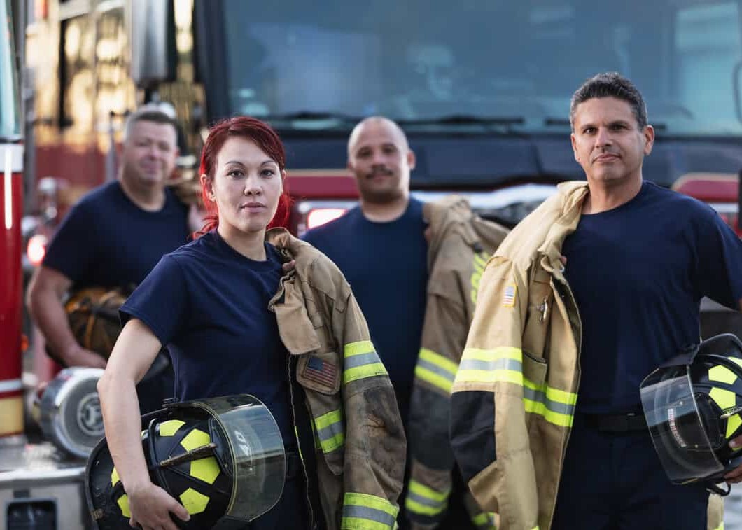 A multiracial group of four firefighters standing in front of two fire engines holding their helmets and fire protection suits, looking at the camera. The focus is on the female firefighter, a Filipino woman in her 30s, in the foreground.