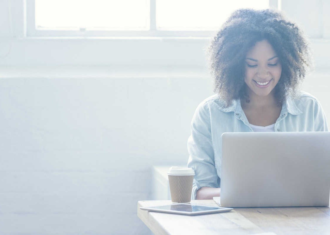 Woman working on a laptop. She is of African descent and has ah afro. There is also a digital tablet and coffee on the table. There is a large window behind her. Copy space