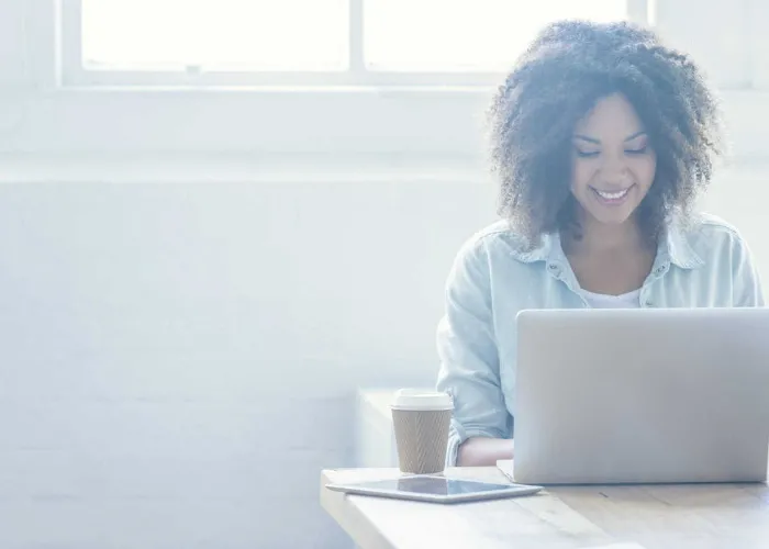 Woman working on a laptop. She is of African descent and has ah afro. There is also a digital tablet and coffee on the table. There is a large window behind her. Copy space